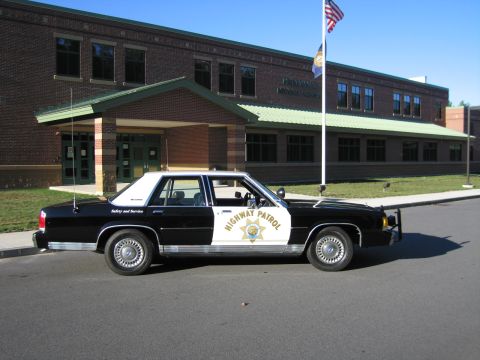 1991 Ford LTD Crown Victoria - Side view of a black-and-white Ford LTD Crown Victoria police sedan with HIGHWAY PATROL door star and roof lights, parked outside a buildin…