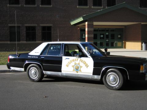 1991 Ford LTD Crown Victoria - Passenger-side profile of a black-and-white Ford LTD Crown Victoria patrol car with gold door star and HIGHWAY PATROL lettering by a buildi…