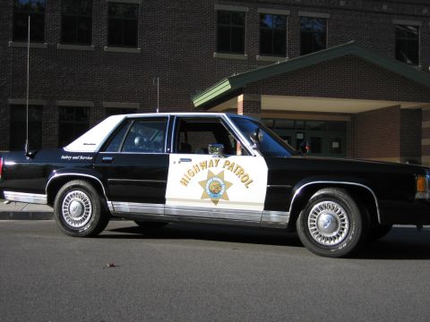 1991 Ford LTD Crown Victoria - Black-and-white Ford LTD Crown Victoria police car in passenger-side profile with gold HIGHWAY PATROL door star, parked by a brick building.