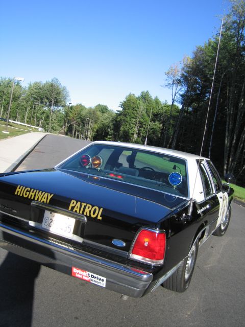 1991 Ford LTD Crown Victoria - Black Ford Crown Victoria police sedan from a low rear three-quarter angle with HIGHWAY PATROL lettering and tall whip antenna on a road.