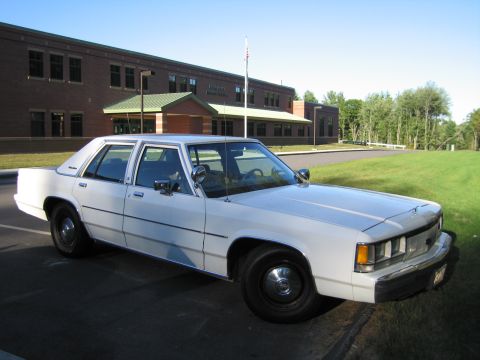 1991 Ford LTD Crown Victoria - white Ford four-door sedan parked on pavement in front of a brick building with a flagpole and lawn.