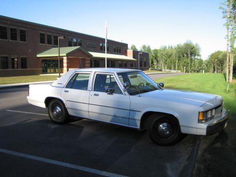 1991 Ford LTD Crown Victoria - Side profile of a white four-door sedan parked in front of a brick building, flagpole, and green lawn.