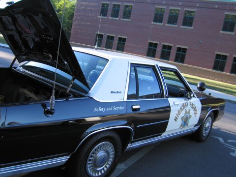 1991 Ford LTD Crown Victoria - black-and-white police sedan marked Highway Patrol and Safety and Service with its hood open beside a brick building.