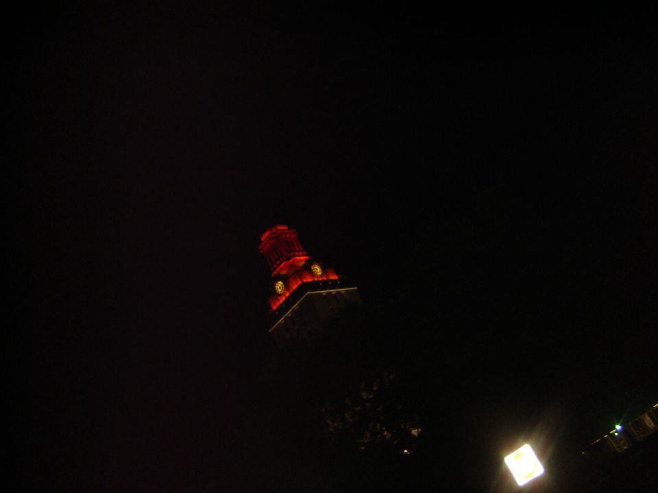 Dark scene with a tall building illuminated by red lights at night. A watermark reads SUPERMOTORS.NET