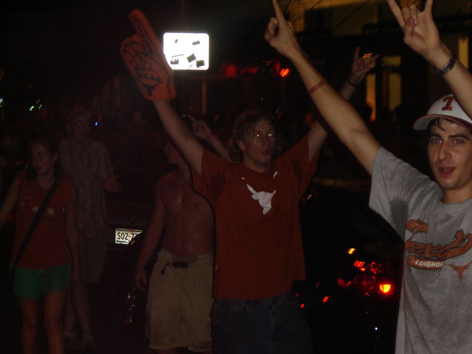 People with arms raised in celebration at night, one holding a large foam finger. A license plate is partially visible