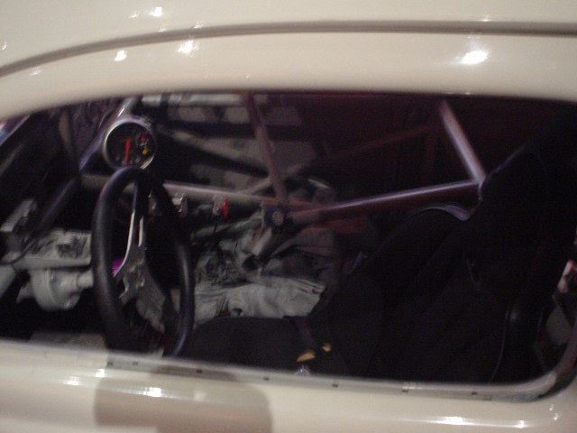 Interior view of a car with a roll cage, racing seat, and a gauge on the dashboard