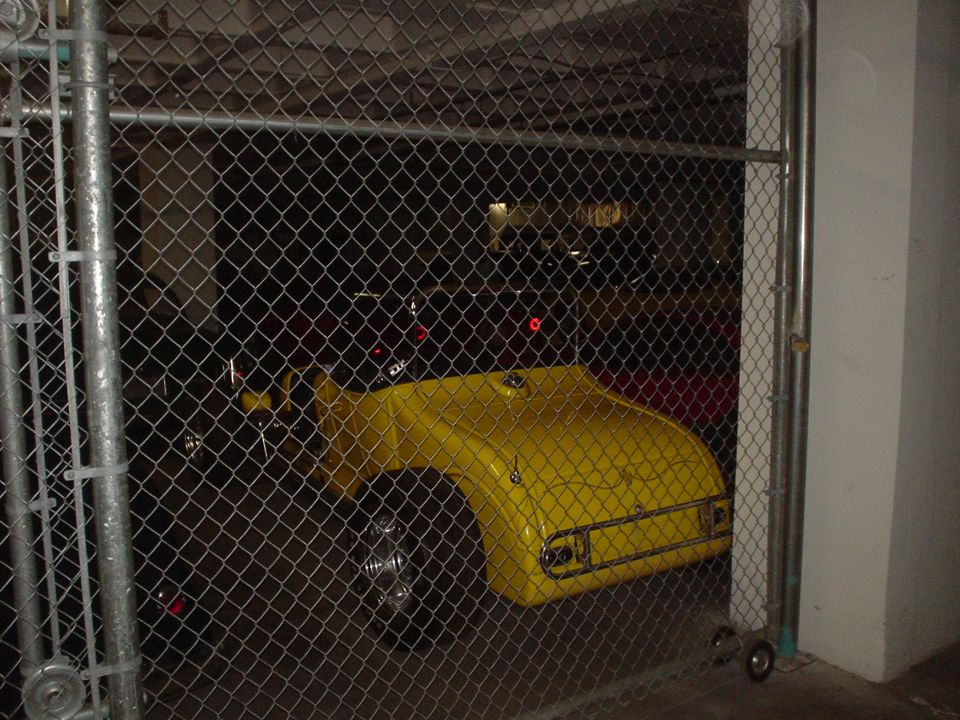 Yellow car body with chrome bumper and taillights, seen through a chain-link fence