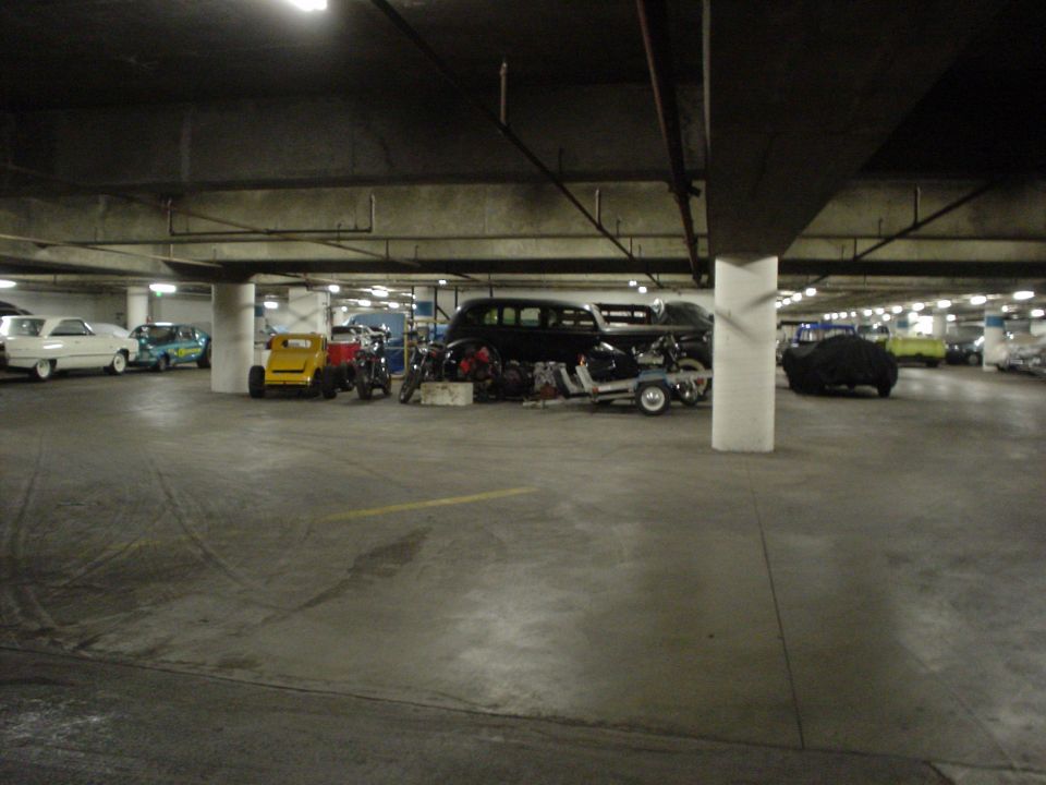 Parking garage scene with a white sedan, a blue car, a yellow go-kart, a black limousine, and a covered vehicle
