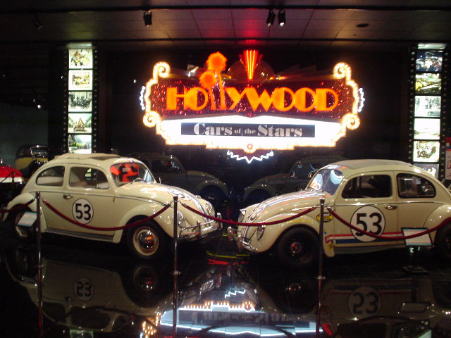 Two white Volkswagen Beetles with number 53 on the side, parked facing each other in front of a 'Hollywood Cars of the Stars' sign
