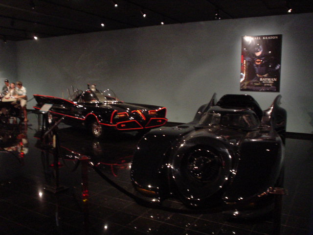 Two black movie cars, one with red trim, on display in a museum with a poster on the wall