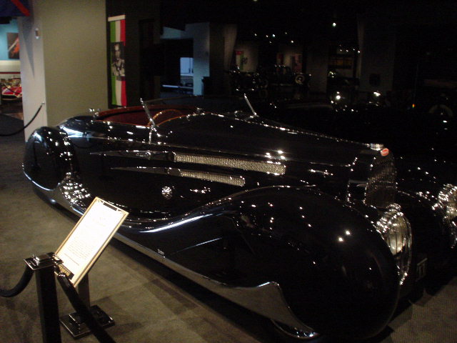 Black vintage convertible with chrome accents and red interior, on display indoors with a descriptive plaque