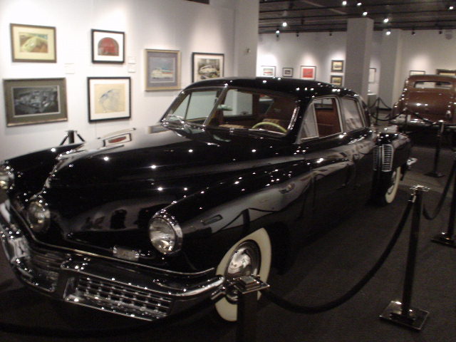 Black sedan with whitewall tires on display in a museum next to other cars and artwork on the wall