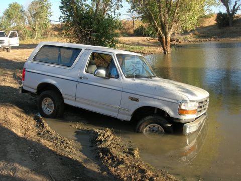 1994 Ford Bronco - A white Ford Bronco is stuck in a muddy body of water, with another vehicle visible in the background.