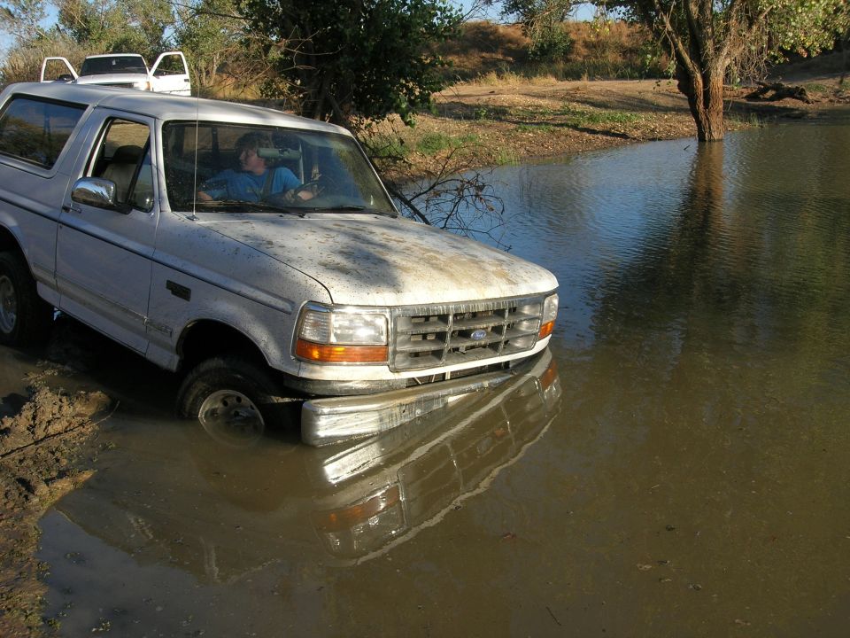 White 1994 Ford Bronco stuck in muddy water, front tires submerged. Another white vehicle visible behind it