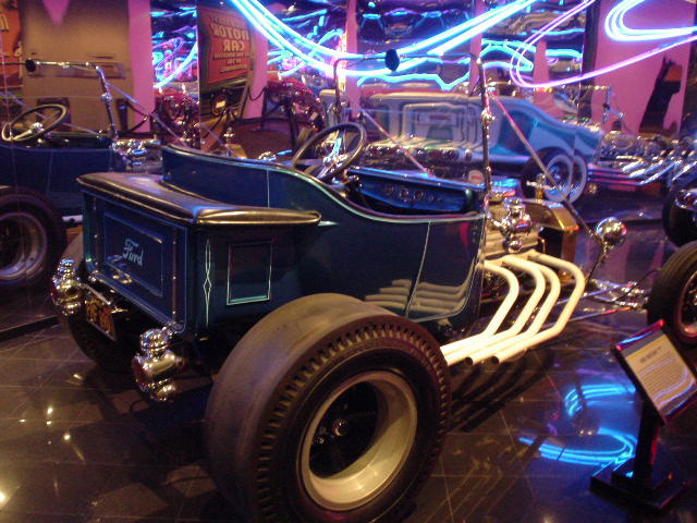 Dark blue hot rod with white exhaust pipes and large rear tires. Another car is visible behind it