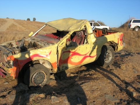 1994 Ford Bronco - A heavily damaged pickup truck sits in a field, showing its flame decals and a flat front tire.