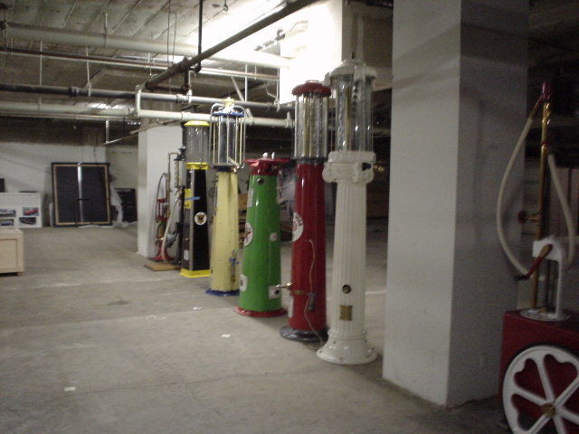 Row of vintage gas pumps in various colors including green, red, yellow, and white, with visible hoses and glass globes