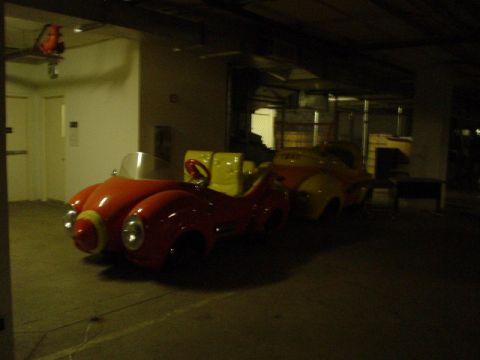 1968 Ford Mustang - Two amusement park ride vehicles, one red and one yellow, are parked indoors in a dimly lit area.