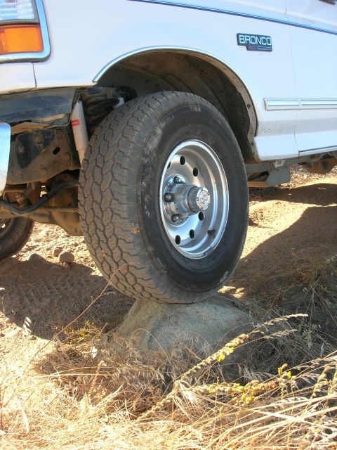 1994 Ford Bronco - A white Ford Bronco's front wheel is positioned on a rock, showing the side profile and a blue 'Bronco' badge.