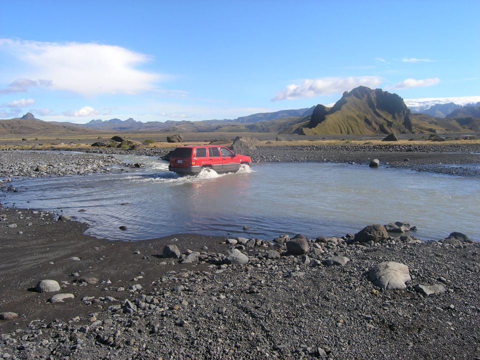 Red SUV driving through a shallow river in a rocky, barren landscape with mountains in the background