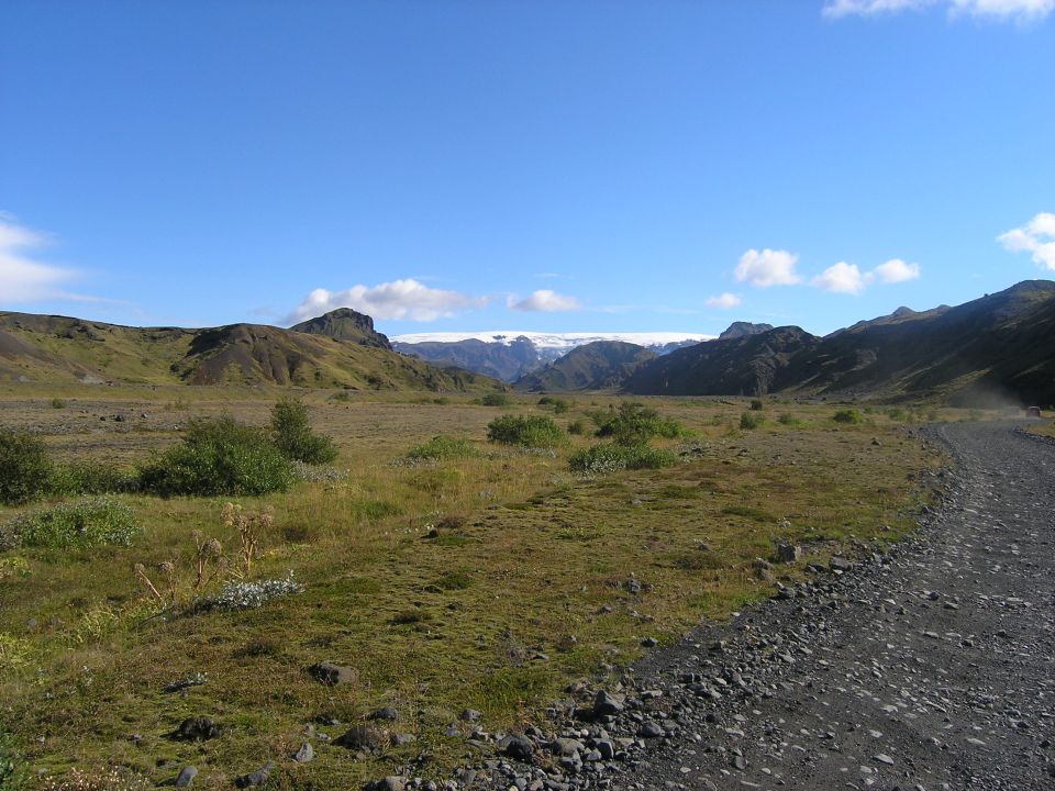 Gravel road leads through a vast, grassy valley towards distant snow-capped mountains under a clear blue sky