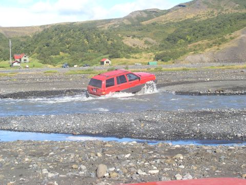 2001 Jeep Wrangler - Red SUV navigates a shallow river in a scenic, mountainous landscape.