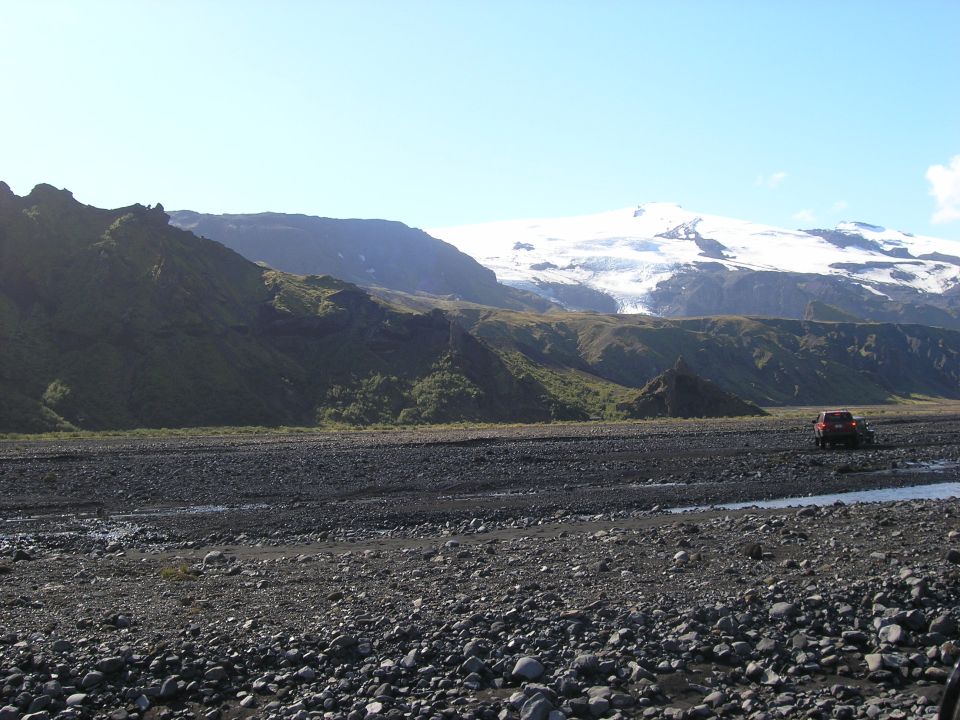 Red SUV driving across a rocky, barren landscape with mountains and a glacier in the background