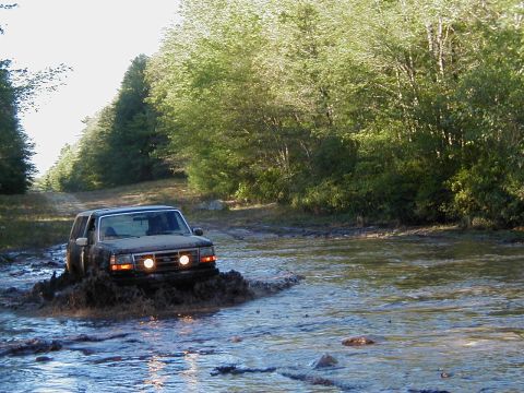1993 Ford Bronco - A mud-caked Ford Bronco pushing a bow wave as it fords a wide, shallow water crossing on a wooded trail, headlights and driving lights on.…