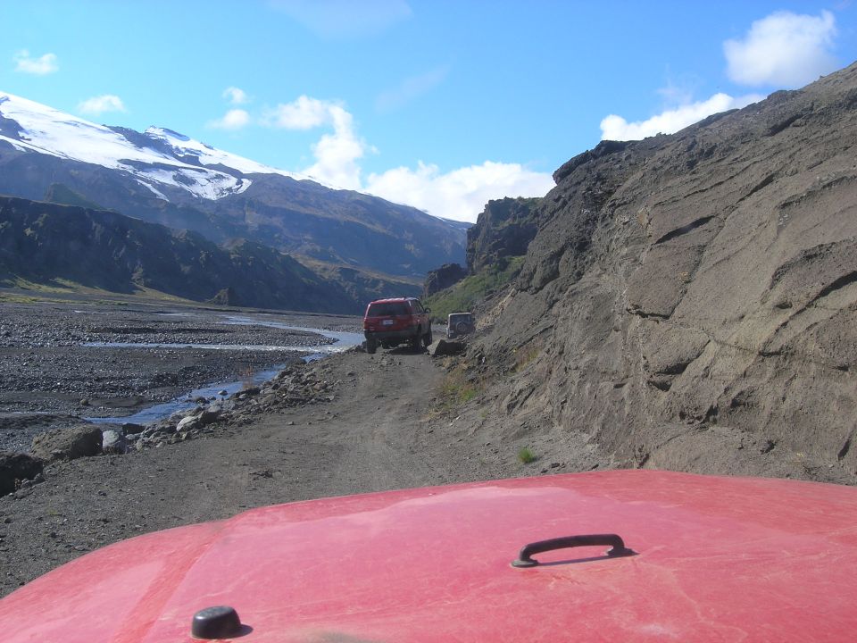 Two red SUVs drive on a dirt road through a rocky valley with a snow-capped mountain in the background
