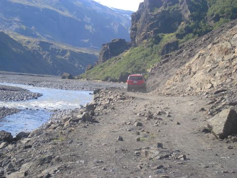 2001 Jeep Wrangler - A red SUV navigates a challenging, rocky path alongside a glacial river in a dramatic mountain landscape.
