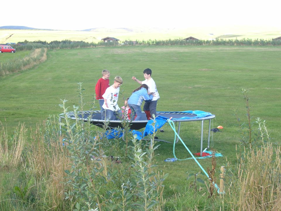 Children jumping on a trampoline in a large grassy field with a red van in the distance