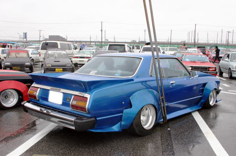 1991 Ford Bronco - A heavily modified blue coupe with a widebody kit and a prominent rear spoiler is seen in a parking lot with other vehicles.