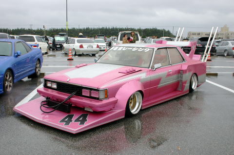 1991 Ford Bronco - A heavily modified pink sedan with custom bodywork and a prominent rear wing sits in a wet parking lot.