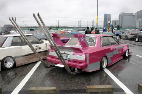1991 Ford Bronco - A heavily modified pink sedan with oversized exhaust pipes and a prominent spoiler is parked next to another customized car.