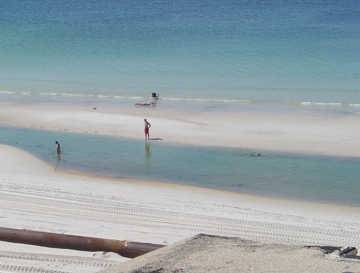 Beach with two people in shallow water and one person standing on shore. Water and sand dominate the scene