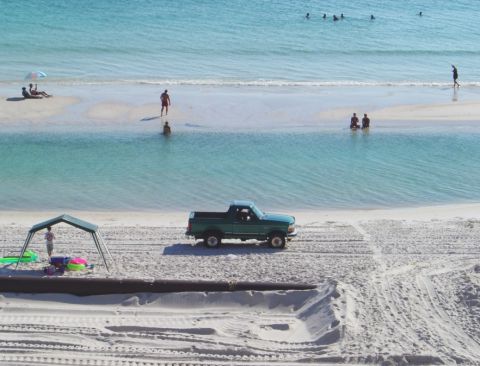 1986 Ford Bronco - Elevated view of a dark green Ford SUV with the roof removed parked on white beach sand beside a tidal lagoon, with swimmers in the turquoi…