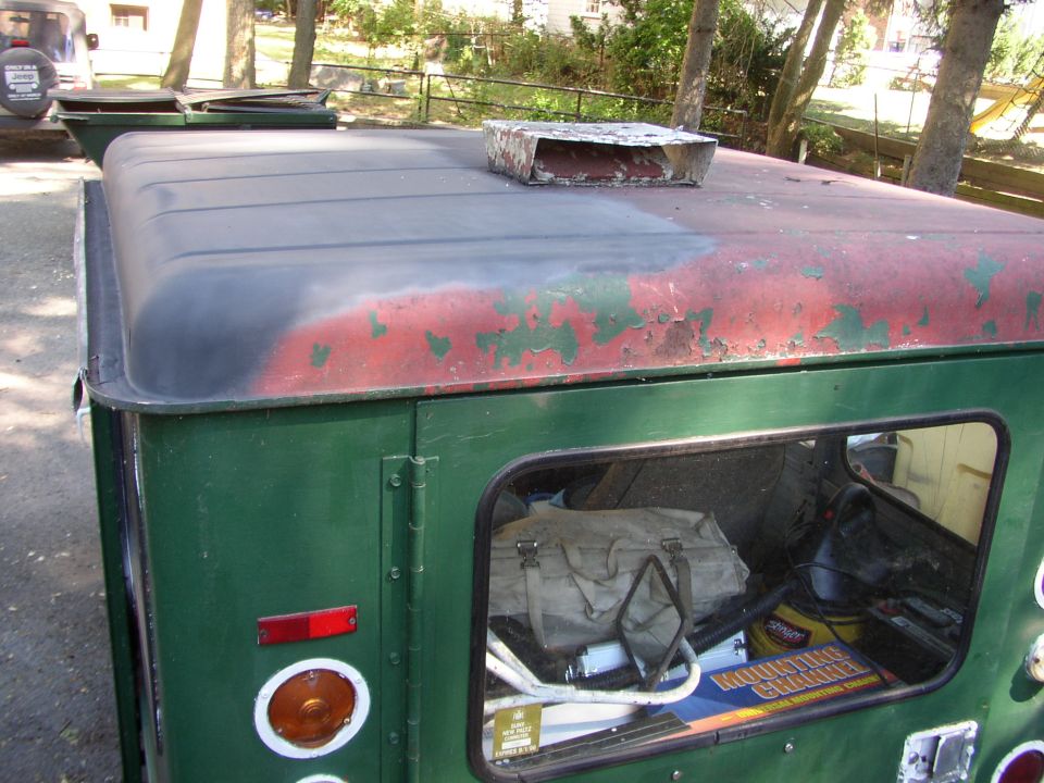 Green Jeep CJ-5 hardtop with peeling red paint on roof, vent on top, and gear inside storage compartment