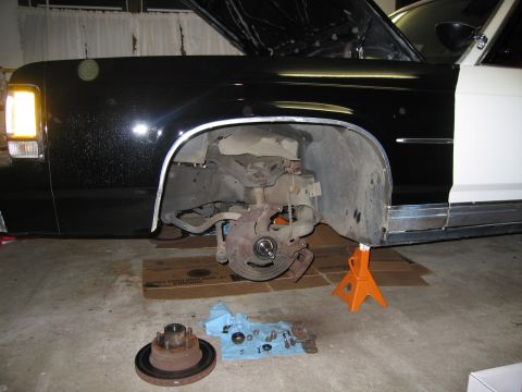 1991 Ford LTD Crown Victoria - Front corner of a black sedan raised on a jack stand with the brake rotor exposed and hub parts on cardboard below.