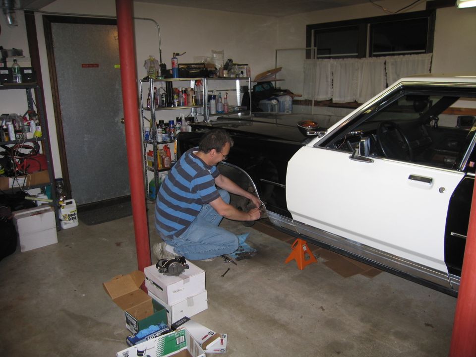 Man in striped shirt kneeling and working on a white sedan in a garage
