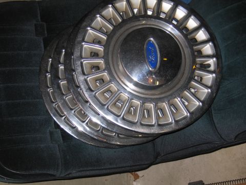 1991 Ford LTD Crown Victoria - Several chrome Ford wheel covers with blue center emblems stacked on a green car seat.
