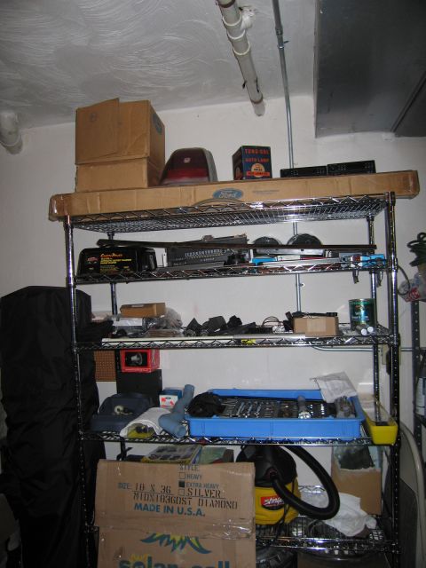 1991 Ford LTD Crown Victoria - metal wire shelving rack against a garage wall holding cardboard boxes, car parts, and assorted electronics.