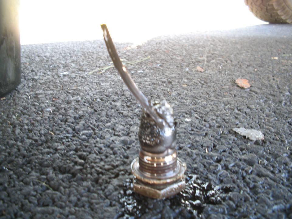 Sludge-covered magnetic drain plug with metal shaving, standing on asphalt beside spilled oil