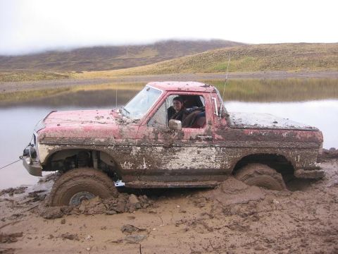 1985 Ford Bronco - A red pickup truck is deeply stuck in mud near a body of water.