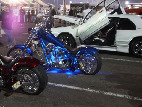 2001 Mercury Grand Marquis - A custom chopper motorcycle with vibrant blue underglow is displayed next to a white car unique gullwing doors at a car show.