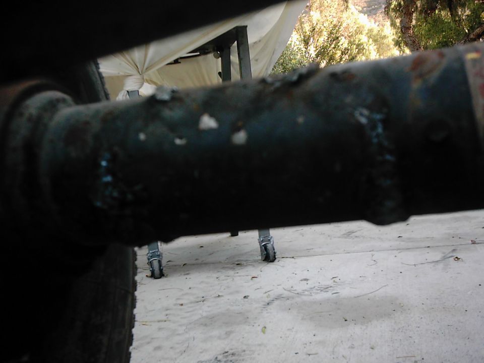 Close-up of a rusted, dark-colored metal pipe, possibly part of a vehicle frame, with small wheels and a white tarp visible in the background