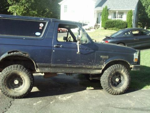 1992 Ford Bronco - This blue Ford Bronco is parked in a driveway next to a house and a dark sedan.