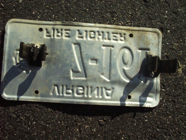 Back of a Virginia Fire Fighter license plate with metal mounting clips, lying on asphalt