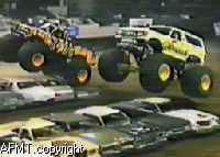 1993 Ford Bronco - Monster trucks perform stunts, jumping over a stack of vehicles in an arena.