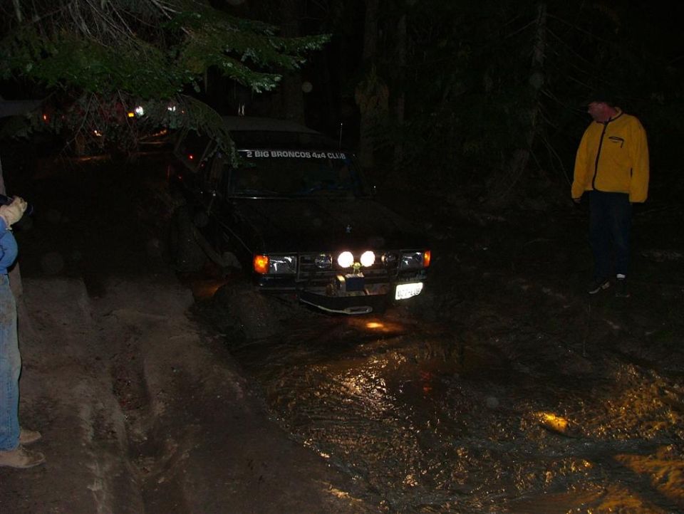 Dark 4x4 truck with lights on entering a muddy trail rut at night as onlookers watch