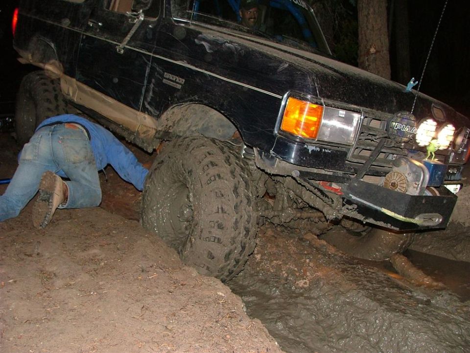 Black lifted Ford Bronco stuck in deep mud at night while a person hooks a strap underneath